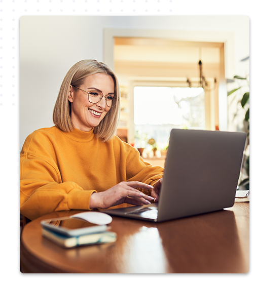 Une femme souriante aux cheveux mi-long, portant des lunettes et un pull orange, est assise à un bureau chez elle, en train de taper sur un ordinateur portable, avec un carnet et un téléphone portable à côté d'elle.
