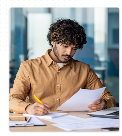 Un jeune homme concentré, portant une chemise marron et avec des cheveux bouclés, examine attentivement des documents de sa formation, tout en prenant des notes avec un stylo jaune, avec un ordinateur portable et d’autres papiers sur sa table de travail.