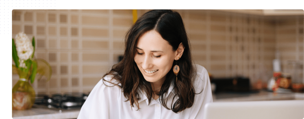 Femme souriante étudiant à distance depuis sa cuisine, carnet et ordinateur devant elle