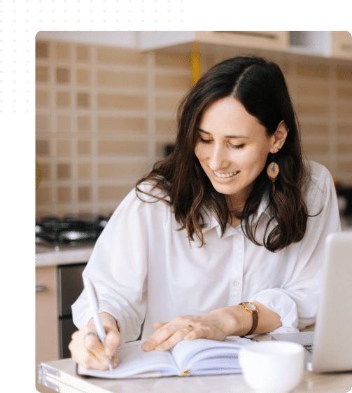 Femme souriante étudiant à distance depuis sa cuisine, carnet et ordinateur devant elle